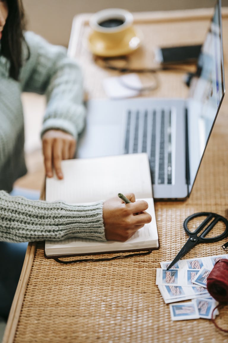 High angle of crop faceless ethnic female student in casual clothes at table in room writing information with pen in notepad near netbook and tea mug near scissors and stamps